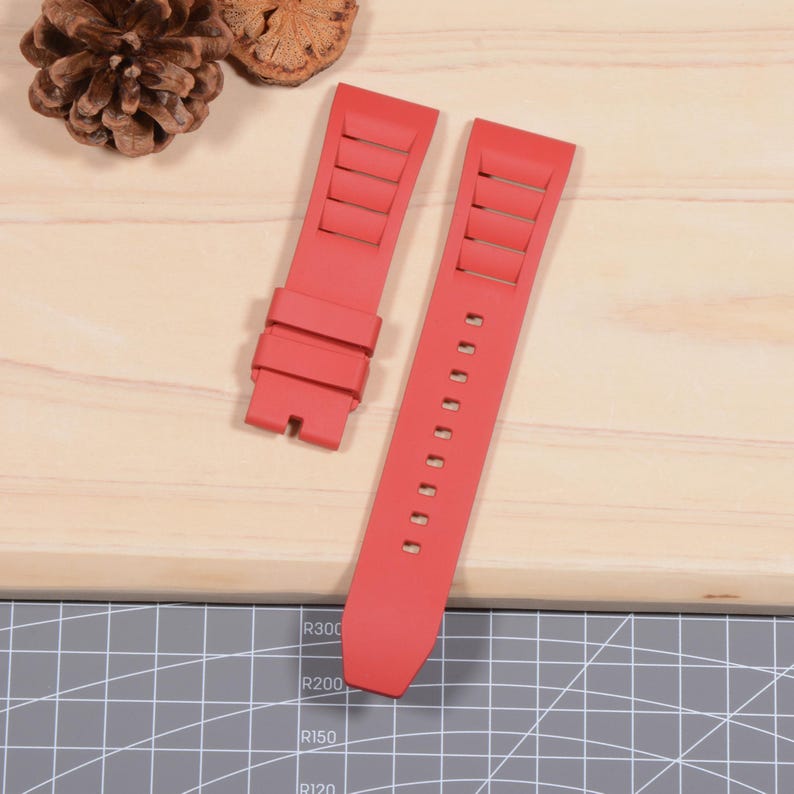 Op de afbeelding: Tweedelig rode horlogeband. De band is gemaakt van een flexibel materiaal en heeft rechthoekige uitsparingen aan &eacute;&eacute;n kant. De andere kant heeft meerdere gaten voor aanpassing. De band wordt getoond op een licht houten oppervlak.