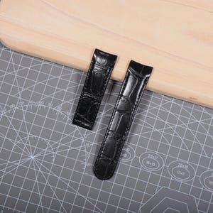 Puede incluir: Dos correas de reloj negras con textura de piel de cocodrilo se muestran sobre una esterilla de corte gris y una superficie de madera clara. Las correas son rectangulares con extremos redondeados.