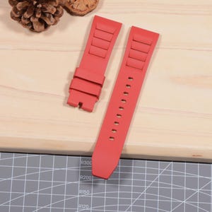 Op de afbeelding: Twee felrode horlogebandjes. E&eacute;n bandje heeft een reeks rechthoekige ribbels, terwijl de andere een reeks kleine gaatjes heeft. De bandjes zijn gemaakt van flexibel materiaal en zijn ontworpen om aan een horloge te bevestigen.