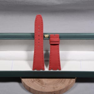 Puede incluir: Correa de reloj roja de dos piezas con superficie texturizada. La correa tiene una serie de agujeros para ajuste y est&aacute; cosida con hilo rojo. La correa del reloj se muestra dentro de una caja verde y blanca.