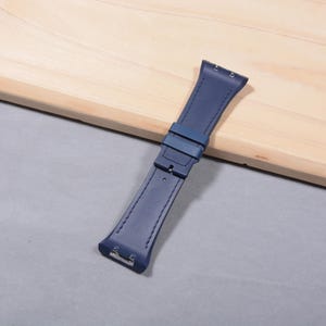 K&ouml;nnte beinhalten: Ein marineblaues Uhrenarmband mit Schnalle und Nahtdetails. Das Armband besteht aus einem glatten Material und wird auf einer hellen Holzoberfl&auml;che gezeigt.
