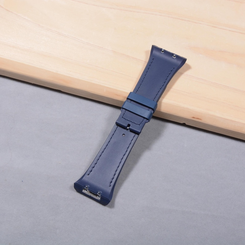K&ouml;nnte beinhalten: Ein marineblaues Uhrenarmband mit Schnalle und Nahtdetails. Das Armband besteht aus einem glatten Material und wird auf einer hellen Holzoberfl&auml;che gezeigt.