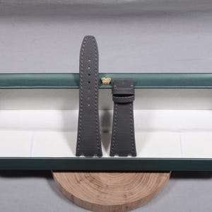 Puede incluir: Dos correas de reloj grises con costuras blancas se muestran dentro de una caja verde con un emblema de corona dorada. Las correas est&aacute;n hechas de un material texturizado. La caja est&aacute; sobre una superficie de madera.