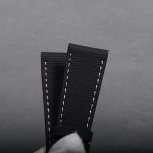 May include: Close-up of a black watch strap with white stitching. The strap is made of a textured material and has a classic design. The background is a solid gray.