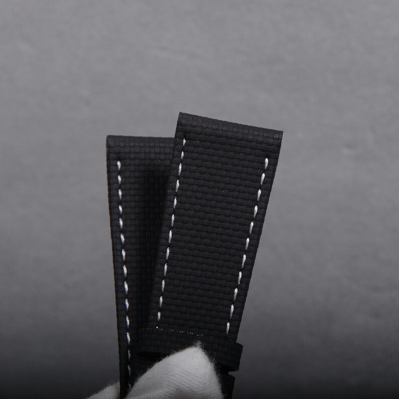 May include: Close-up of a black watch strap with white stitching. The strap is made of a textured material and has a classic design. The background is a solid gray.