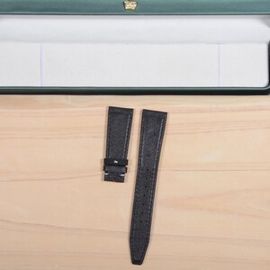 May include: Two black leather watch straps with a textured finish. One strap has a buckle, and the other has multiple holes for adjustment. The straps are displayed on a light wooden surface, with a green box in the background.