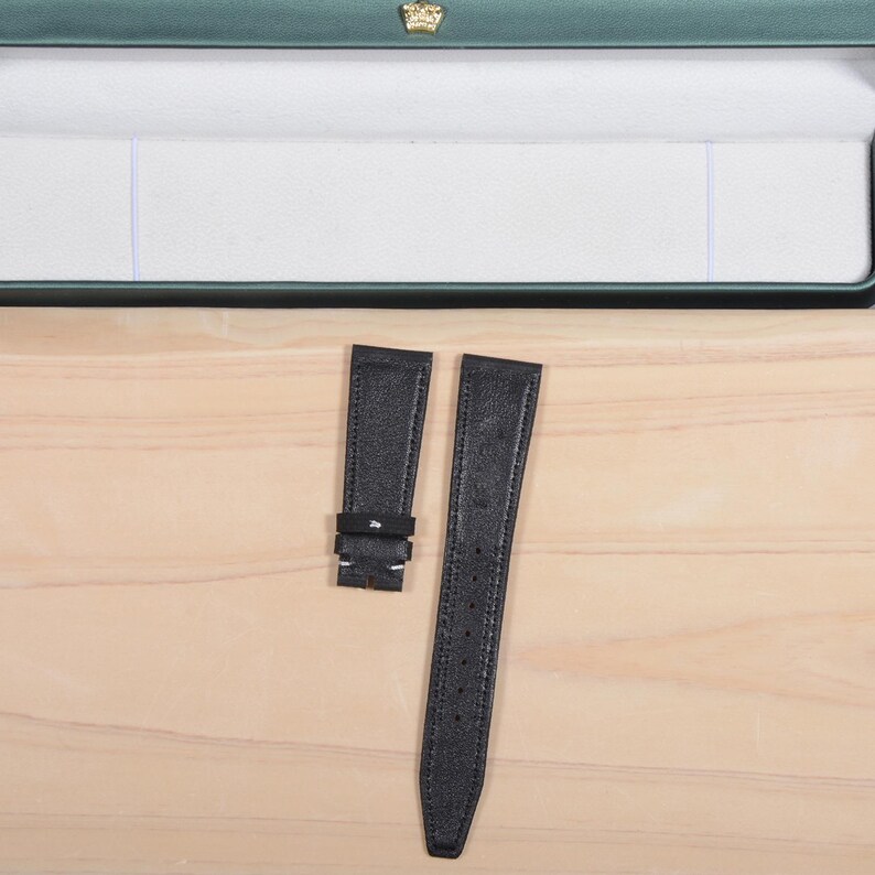 May include: Two black leather watch straps with a textured finish. One strap has a buckle, and the other has multiple holes for adjustment. The straps are displayed on a light wooden surface, with a green box in the background.