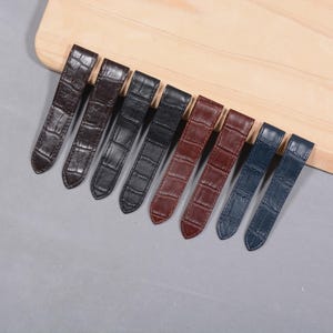 Könnte beinhalten: Eine Sammlung von acht Uhrenarmbändern in verschiedenen Farben, darunter Braun, Schwarz und Blau. Die Armbänder haben ein strukturiertes, krokodilartiges Muster und sind auf einer hellen Holzoberfläche angeordnet.
