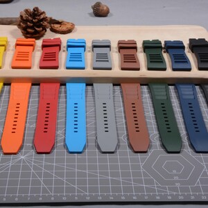 Op de afbeelding: Een verzameling kleurrijke horlogebandjes in diverse tinten, waaronder oranje, rood, blauw, grijs, bruin, groen en zwart. De bandjes worden getoond op een houten oppervlak en een grijze snijmat.