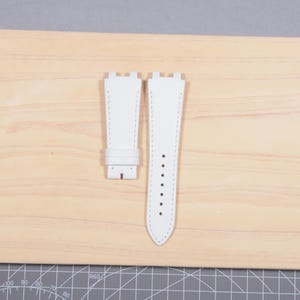 May include: A pair of white leather watch straps with white stitching. The straps are shown on a light wood surface. One strap has a buckle, and the other has several holes for adjustment. The straps are new and unused.