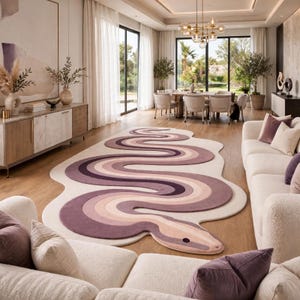 Puede incluir: Una alfombra grande de color crema con un diseño prominente y sinuoso en forma de serpiente en varios tonos de morado, malva y rosa suave. La alfombra tiene una forma orgánica irregular que sigue las curvas del patrón de serpiente.