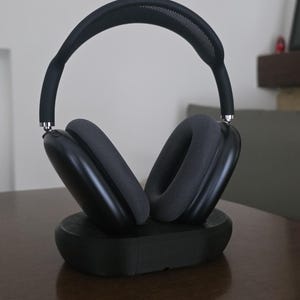 May include: Black over-ear headphones with dark grey ear cushions and headband, on a black charging base. The headphones are on a brown wooden table. A framed artwork hangs in the background.