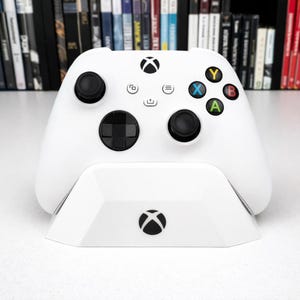 Puede incluir: Un mando y una base de carga Xbox blancos. El mando tiene joysticks y botones negros con detalles de colores. La base presenta el logotipo de Xbox.