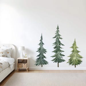 May include: Watercolor-style wall decals of three evergreen trees in varying shades of green. The trees are affixed to a white wall, creating a nature-inspired accent. The room also features a bed, nightstand, lamp, and rug.