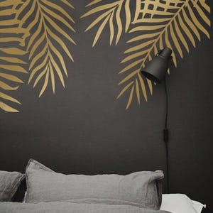 May include: A bedroom scene featuring a dark gray wall adorned with gold palm leaf decals. A black adjustable lamp is mounted on the wall. The bed is covered with gray bedding and pillows, creating a modern and tropical aesthetic.