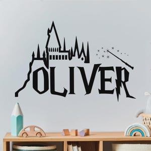 May include: Black wall decal featuring the name "OLIVER" in a stylized font, with a castle silhouette, a wand, and stars. The design is suitable for a child's room or nursery.