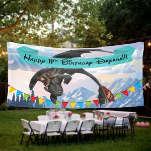 May include: A large banner with a cartoon dragon and the text "Happy 11th Birthday Dapana!!!" is displayed outdoors. The banner has a blue sky and mountain background, with colorful pennant flags below. A party table is set up below the banner.