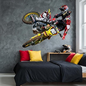 Peut inclure: Une décoration murale dynamique représentant un pilote de motocross en plein vol sur une moto Suzuki jaune et noire. Le pilote porte une combinaison et un casque de course rouges, noirs et blancs. La scène se déroule sur un mur gris texturé au-dessus d'un lit avec une literie noire et des coussins colorés.
