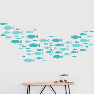 Pode incluir: Um decalque de parede com um cardume de peixes estilizados em tons de azul e verde-azulado. Os peixes estão dispostos em um padrão ondulado. O fundo é uma parede branca lisa.
