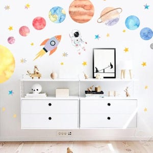 May include: A whimsical space-themed wall decal set featuring planets, stars, a rocket, an astronaut, and a UFO. The decals are in watercolor style and are arranged on a white wall above a white storage unit and a road-themed rug.