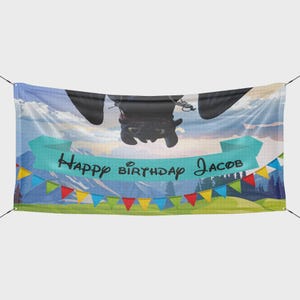 May include: A banner featuring a cartoon dragon hanging upside down, with the text "Happy Birthday Jacob" on a teal banner. The background shows a mountain landscape with colorful bunting.