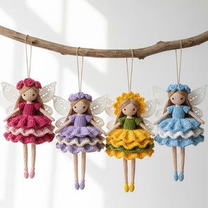 Op de afbeelding: Vier handgemaakte gehaakte feeënpoppen hangen aan een houten tak. Elke pop heeft een jurk in een andere kleur: roze, paars, geel en blauw. Ze hebben gehaakte vleugels en bloemenkransen. De poppen hangen aan touwtjes.