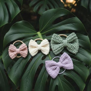 May include: Four handmade crochet hair bows in pastel colors: pink, cream, green, and lavender. Each bow is attached to a neutral-toned elastic hair tie. The bows are displayed on a large, green tropical leaf.