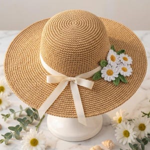 May include: A beige crocheted sun hat with a wide brim and a cream-colored ribbon tied in a bow. The hat is adorned with a cluster of white and yellow crocheted daisies and green leaves. The hat is displayed on a white pedestal.