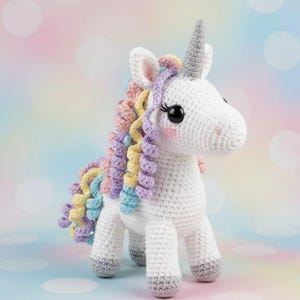 Könnte beinhalten: Gehäkeltes Einhorn-Spielzeug mit weißem Körper, grauen Hufen und einem silbernen Horn. Die Mähne des Einhorns ist bunt in Gelb-, Rosa-, Lila- und Blautönen gehalten. Das Spielzeug hat große schwarze Augen und rosige Wangen, vor einem pastellfarbenen Hintergrund.