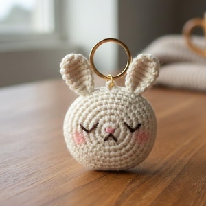 May include: A crocheted bunny keychain with a gold-colored ring. The bunny is cream-colored with closed eyes, pink cheeks, and a pink nose. The keychain is on a wooden surface.