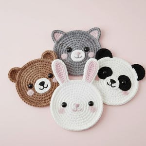 Puede incluir: Cuatro posavasos de ganchillo hechos a mano con forma de animales: un oso pardo, un gato gris, un conejo blanco y un oso panda. Cada posavasos presenta detalles bordados, incluyendo ojos, narices y rubor. Los posavasos están dispuestos sobre una superficie rosa.