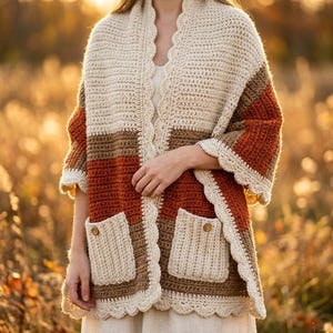 May include: A handmade crochet shawl in cream, brown, and rust-orange hues. The shawl features a scalloped edge, two front pockets, and button details. The garment is worn over a white dress, with a blurred background of tall grass.
