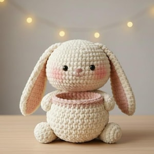 Könnte beinhalten: Ein handgefertigter, cremefarbener Häschenkorb mit rosa Akzenten. Das Häschen hat lange Ohren mit rosa Innenohren, rosigen Wangen und schwarzen Augen. Der Korb ist rund mit einem rosa Rand und steht auf einer Holzoberfläche.