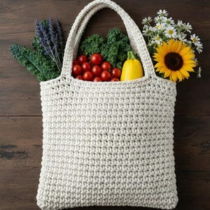 Peut inclure: Un sac fourre-tout en crochet de couleur crème rempli de produits frais et de fleurs. Le sac contient du chou frisé, des tomates cerises, une courge jaune, de la lavande, un tournesol et des marguerites. Le sac est posé sur une surface en bois foncé.