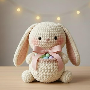 Może przedstawiać: Szydełkowy królik z koszykiem wielkanocnych jajek. Królik jest kremowy z różowymi akcentami na uszach i policzkach. Różowa kokarda zdobi przód. Koszyk zawiera małe, pastelowe jajka.