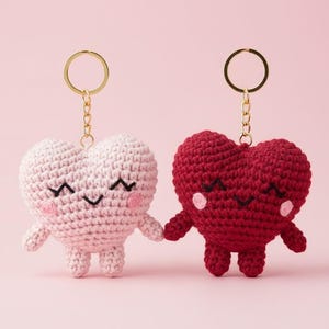 May include: Two crochet heart-shaped keyrings. One is light pink, the other is red. Both have smiling faces, rosy cheeks, and small arms and legs. Each keyring has a gold-coloured ring.