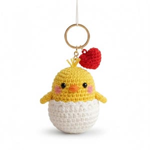 May include: A handmade crochet keychain featuring a yellow chick emerging from a white eggshell. The chick has black eyes, pink cheeks, and an orange beak. A small red heart charm hangs above the chick, attached to a gold-colored keyring.