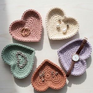 Könnte beinhalten: Eine Sammlung von sechs herzförmigen Häkeltabletts in Pastellfarben wie Rosa, Creme, Grün, Braun und Lavendel. Jedes Tablett enthält Schmuck und eine Uhr. Die Tabletts sind kreisförmig auf einer hellen Oberfläche angeordnet.