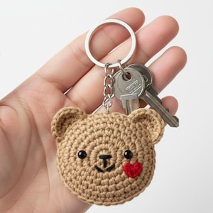 Puede incluir: Llavero de oso de peluche tejido a ganchillo en color beige con ojos negros y un corazón rojo. El llavero está unido a un anillo plateado con dos llaves.