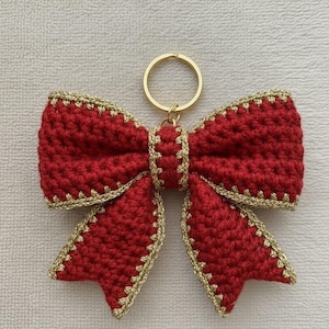 May include: A red crochet bow keyring with gold edging and a gold-coloured key ring. The bow is a decorative item, suitable for adding a handmade touch to keys, bags, or other belongings.
