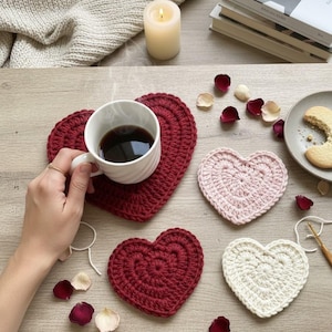 May include: A cozy scene featuring heart-shaped crochet coasters in red, pink, and white. A white mug filled with coffee sits on a red coaster. Rose petals and a lit candle add to the warm aesthetic. A plate with a cookie is also present.