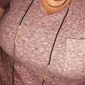 May include: A long layered necklace with black and gold beads. The necklace is worn by a person wearing a maroon shirt.