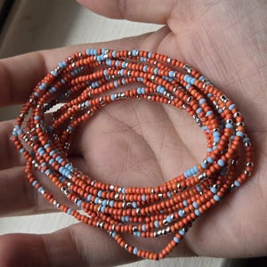 May include: Strands of orange, light blue, and silver beaded jewelry. The beads are small and strung together, forming multiple layers. The jewelry is held in a hand, showcasing the colors and textures.