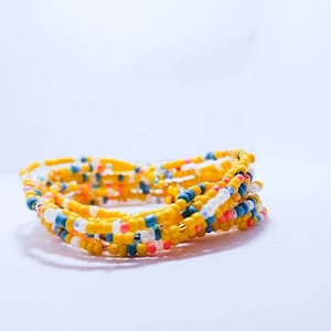 May include: A close-up of a yellow beaded bracelet with blue, white, and pink beads. The bracelet is coiled up on a white surface.