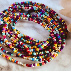 May include: Multicolored beaded waist beads with gold, silver, and white beads. The beads are arranged in a circular pattern and are strung on a thin cord.