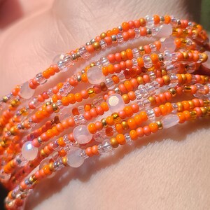 May include: A beaded bracelet wraps around a wrist. The bracelet features orange, gold, and clear beads, with larger, pale pink beads interspersed throughout. The beads are strung together, creating a layered effect. The bracelet is a vibrant accessory.