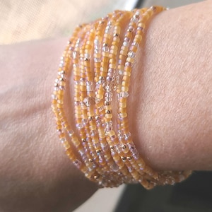 May include: A multi-strand bracelet made of small, iridescent beads in shades of gold, white, and pink. The bracelet is wrapped around a person's wrist.