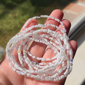 May include: A white beaded waist beads with silver and iridescent accents. The beads are strung together in a circular shape and are held in a hand.