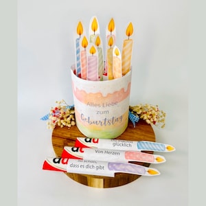 Op de afbeelding: Een verjaardags-thema middelpunt met een cilindrische container met pastelkleurige lagen en papieren kaarsen. De container heeft de tekst "Alles Liebe zum Geburtstag". Extra kaarten met tekst zijn inbegrepen.
