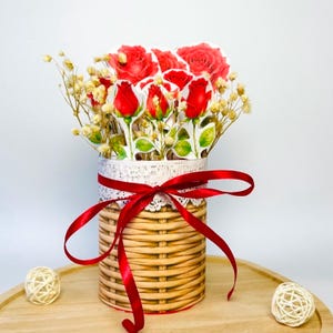 May include: A decorative floral arrangement featuring red paper roses and dried flowers in a woven basket-style container. The container is adorned with a white lace trim and a red ribbon tied in a bow. The arrangement sits on a wooden tray.
